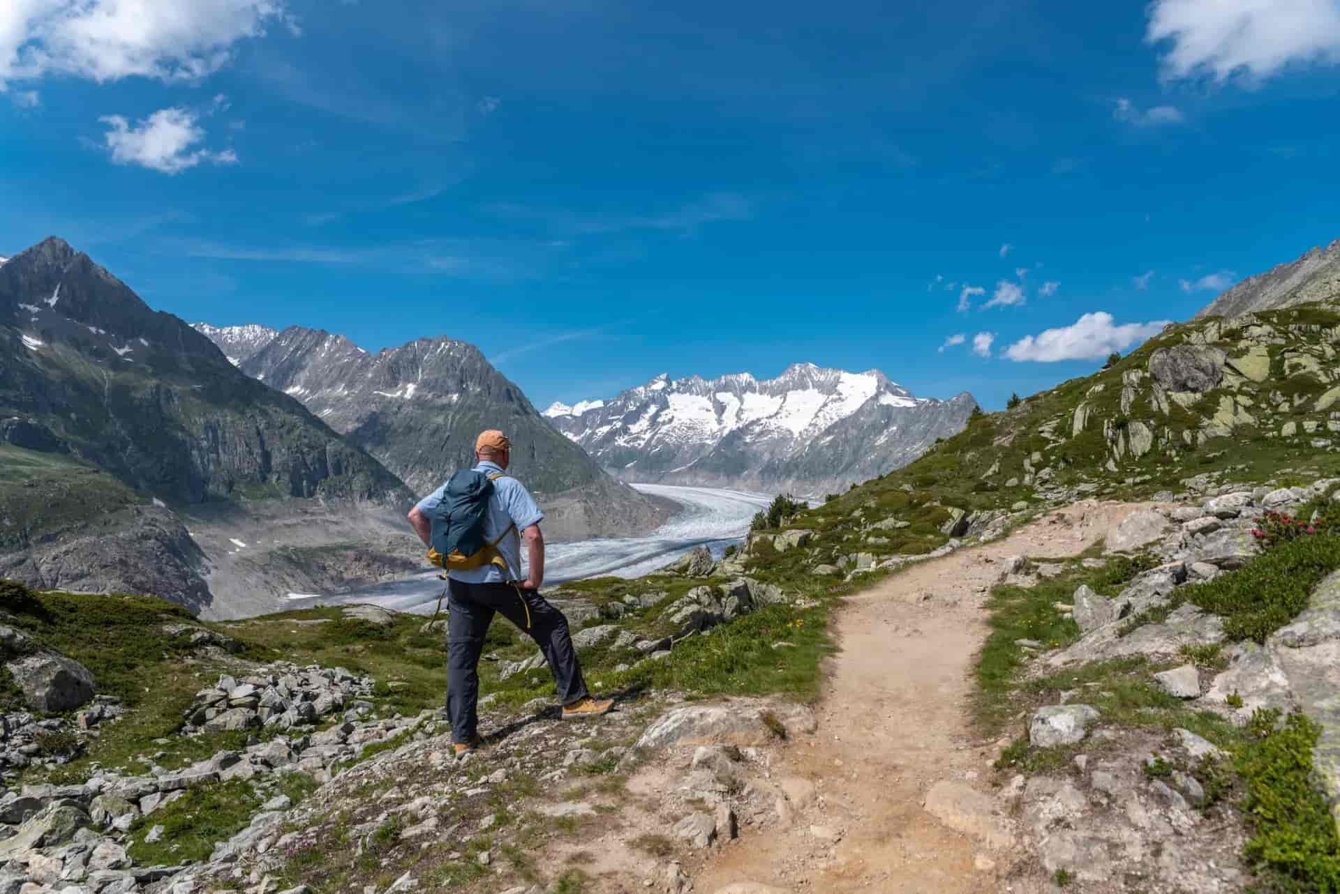 Hiker overlooking Aletsch Glacier Panorama Trail with snow-capped mountains under blue sky