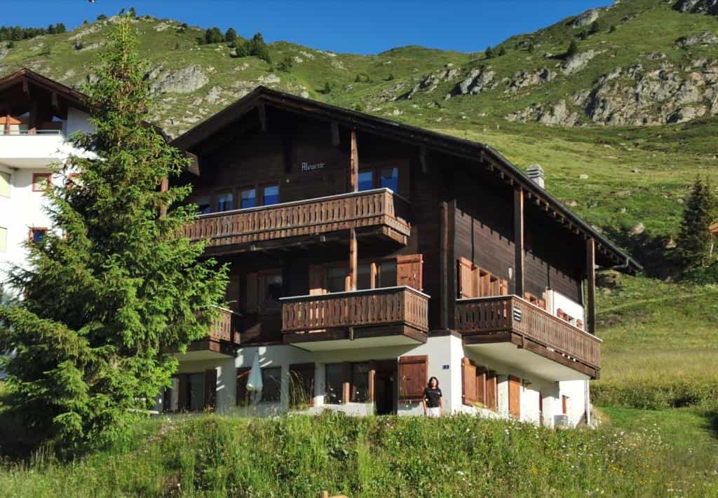Wooden chalet in Riederalp against a green, rocky mountainside under a blue sky.