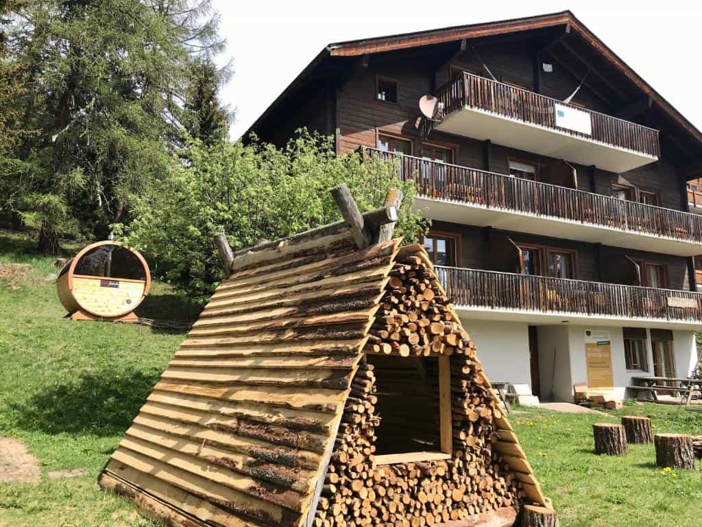 Wooden log storage structure, barrel sauna, and chalet in a grassy alpine setting