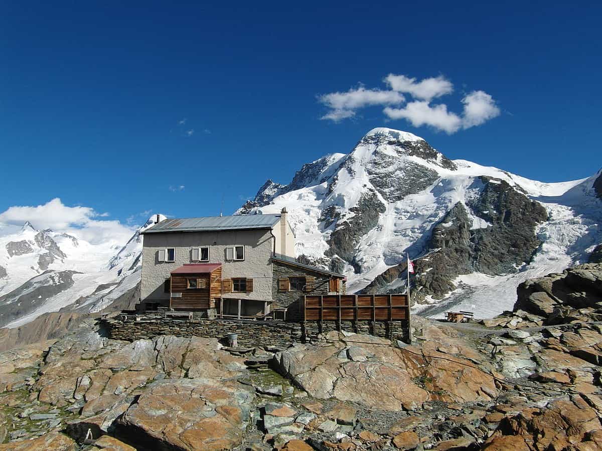 Alpine hut on rocky terrain below massive snow-covered mountain peaks under a blue sky.