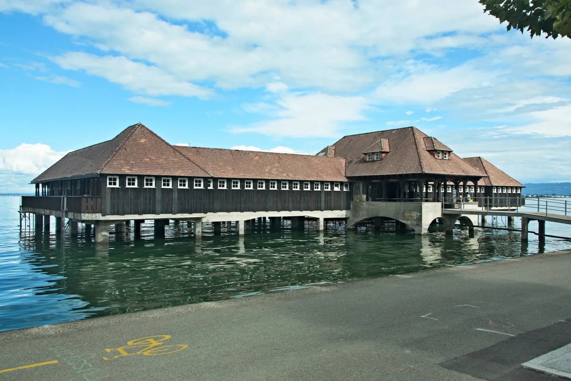 Historic wooden bath house on stilts over lake water with bicycle symbol on pavement.