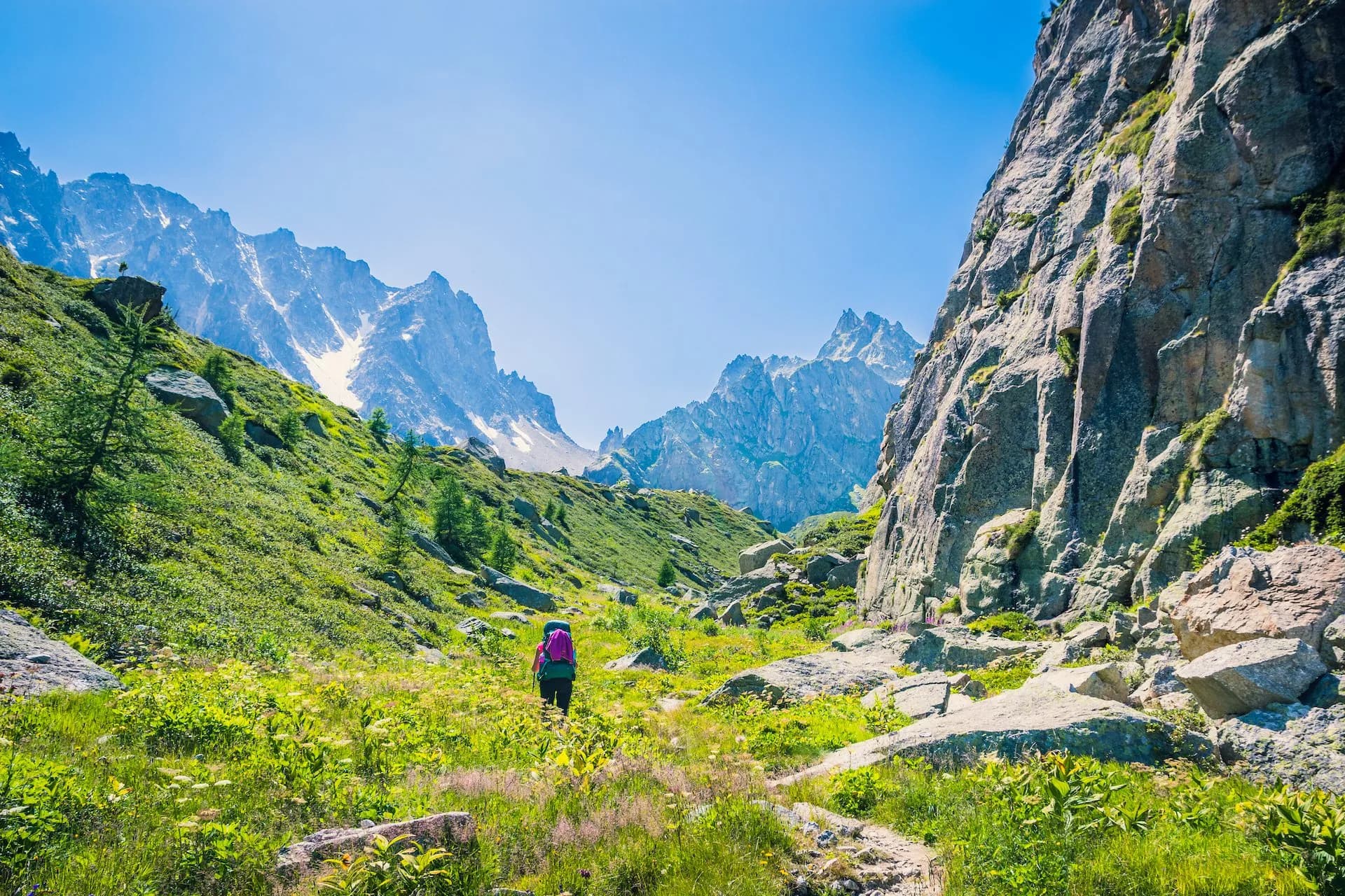 Hiker with backpack walking through lush green alpine valley toward jagged, snow-dusted mountains.