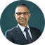 Portrait of a man wearing glasses, suit, and tie against a dark teal background.