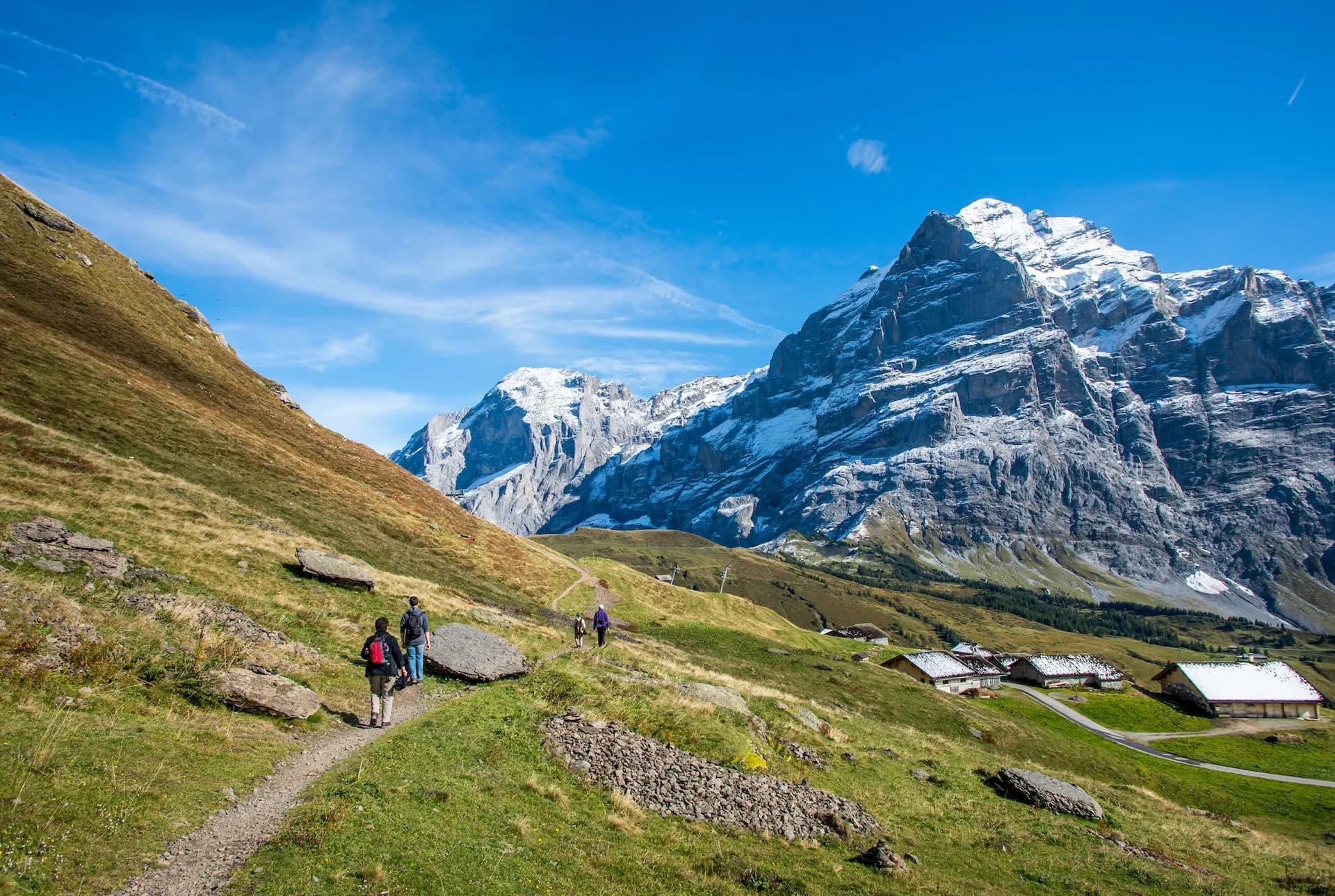 Hikers on a trail toward Grosse Scheidegg with snow-capped alpine mountains under a blue sky.