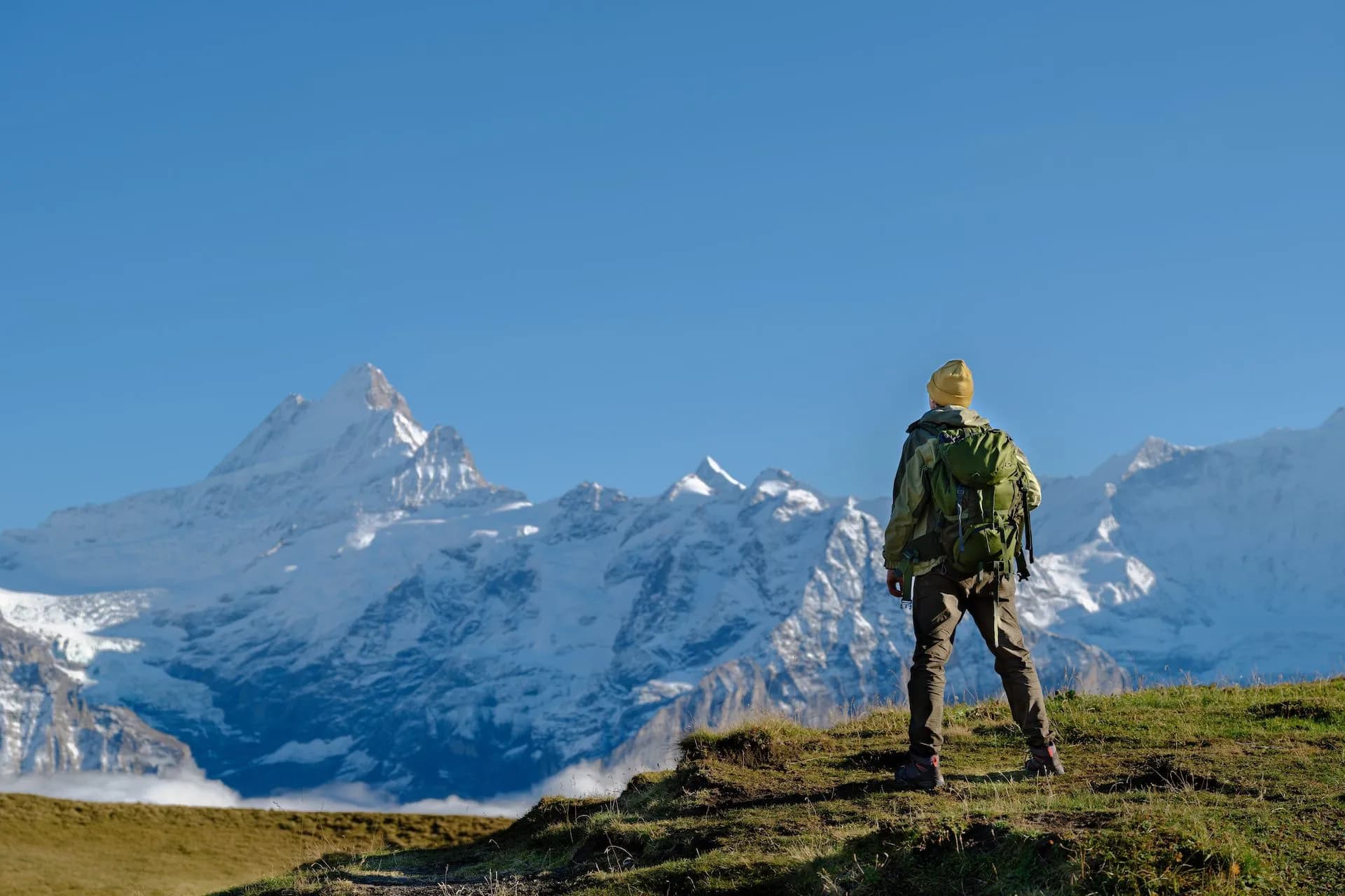 Hiker with backpack standing on grassy hill facing snow-covered mountains above Grindelwald.