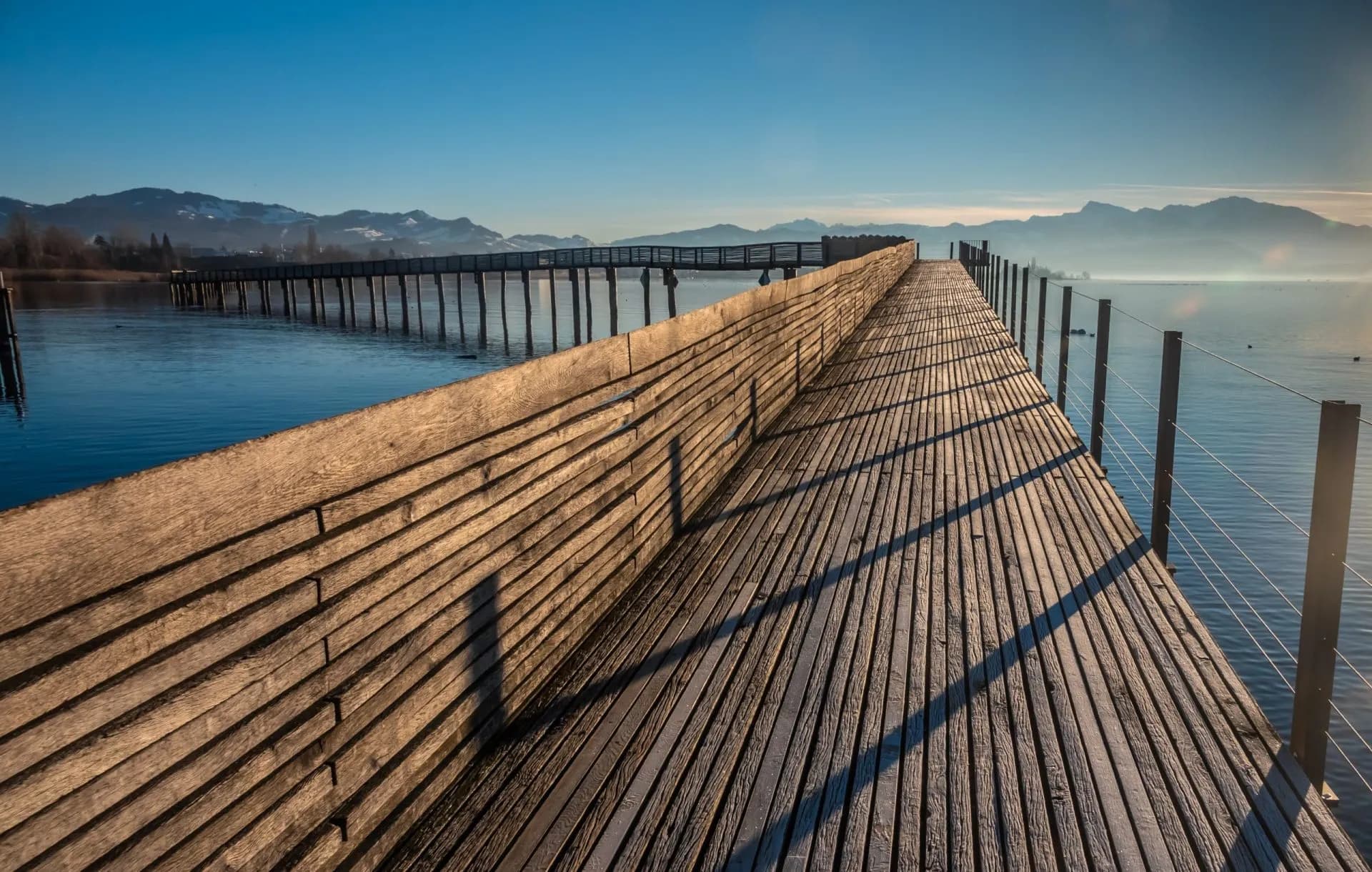 Long wooden footbridge over Lake Zurich with distant mountains under a clear blue sky.