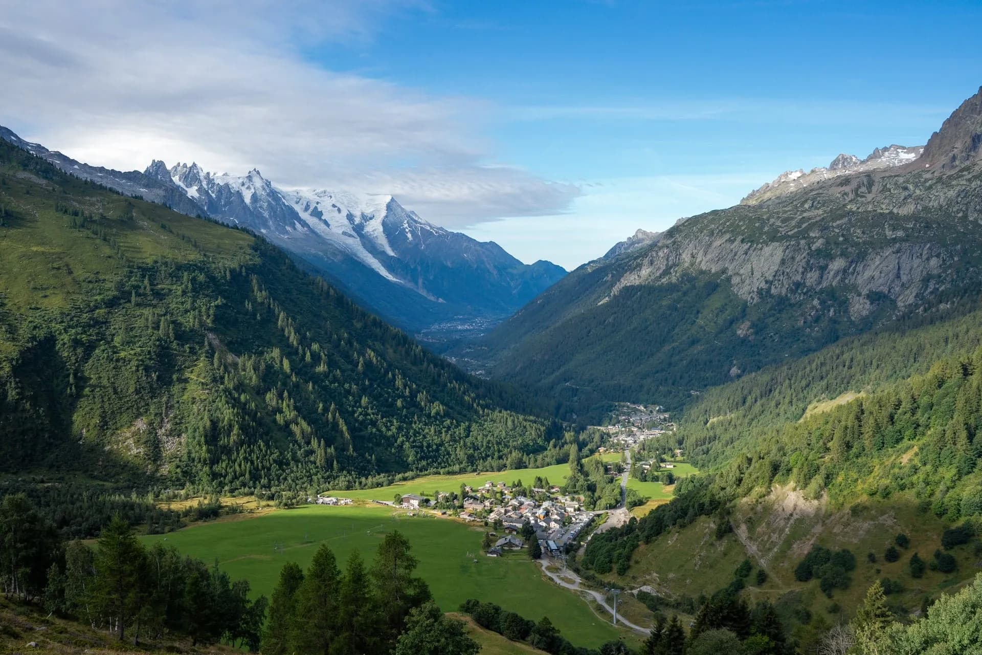 Alpine valley with forested slopes, green meadows, a small village, and snow-capped mountains in Chamonix Valley.