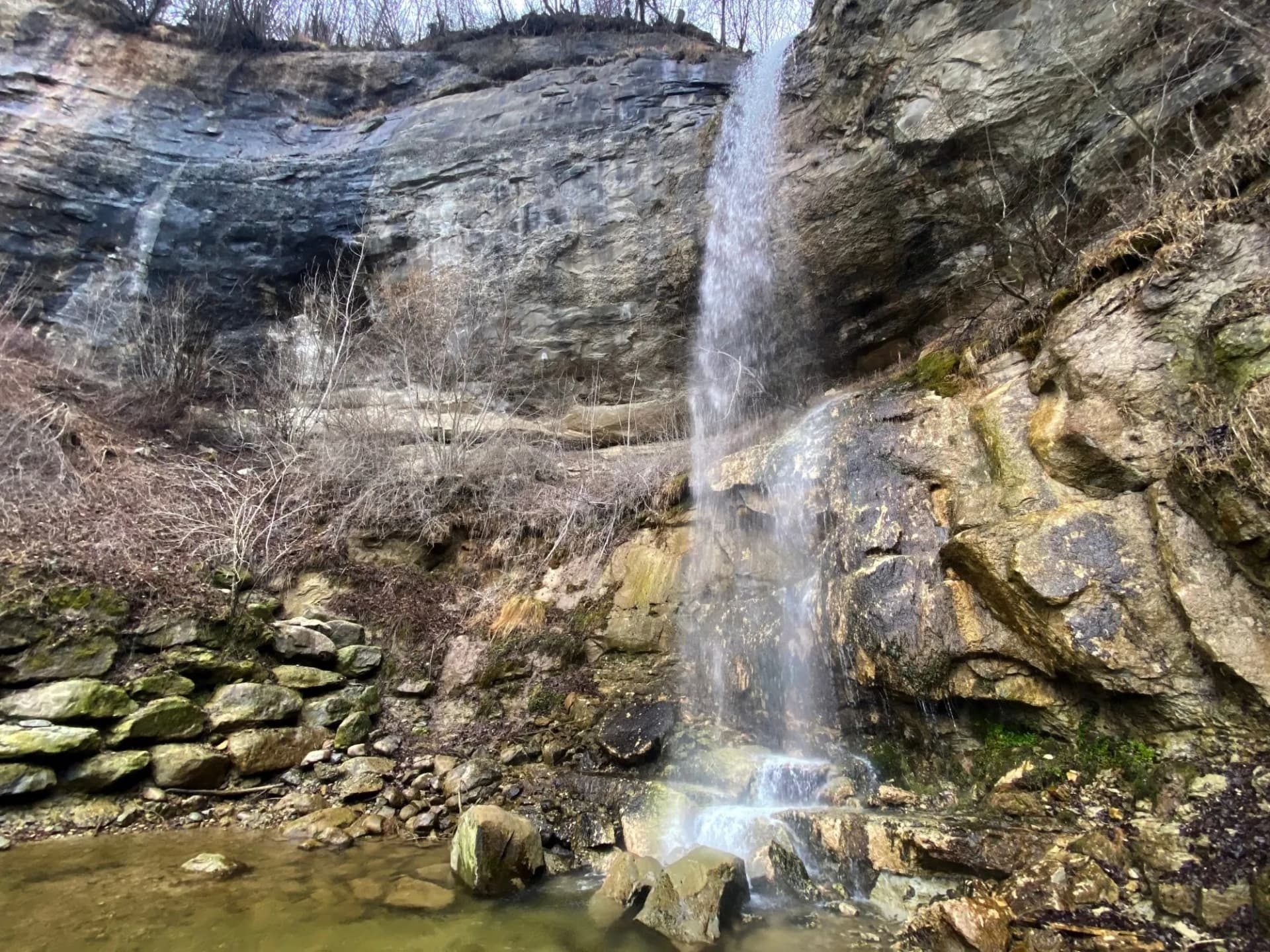 Waterfall cascading down a rocky cliff face into a shallow pool with bare winter brush.