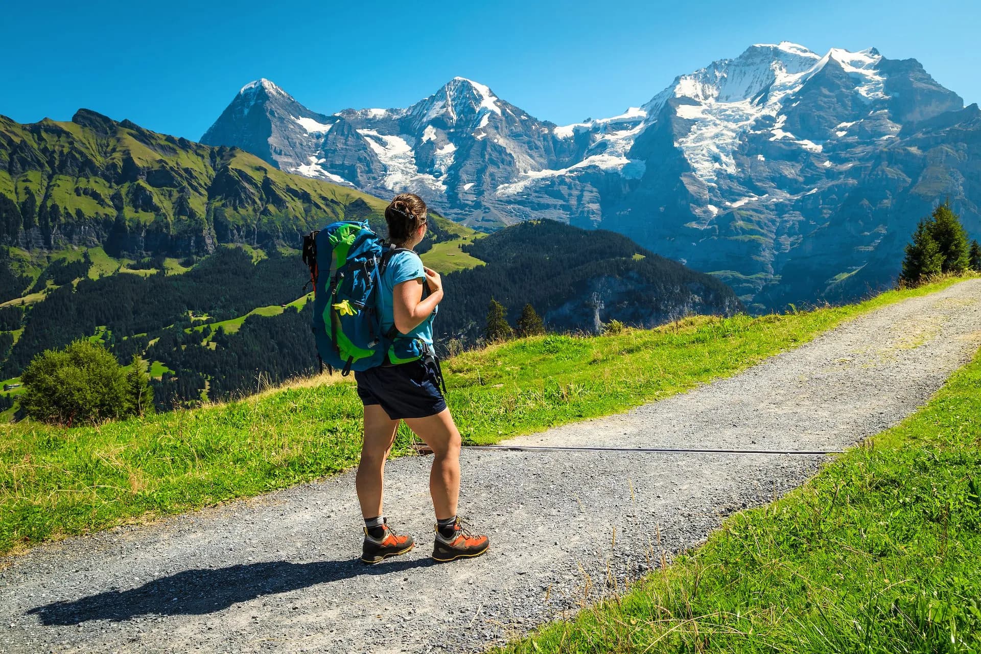 Hiker with backpack on gravel path overlooking snow-capped mountains on the Mürren Mountain View Trail.