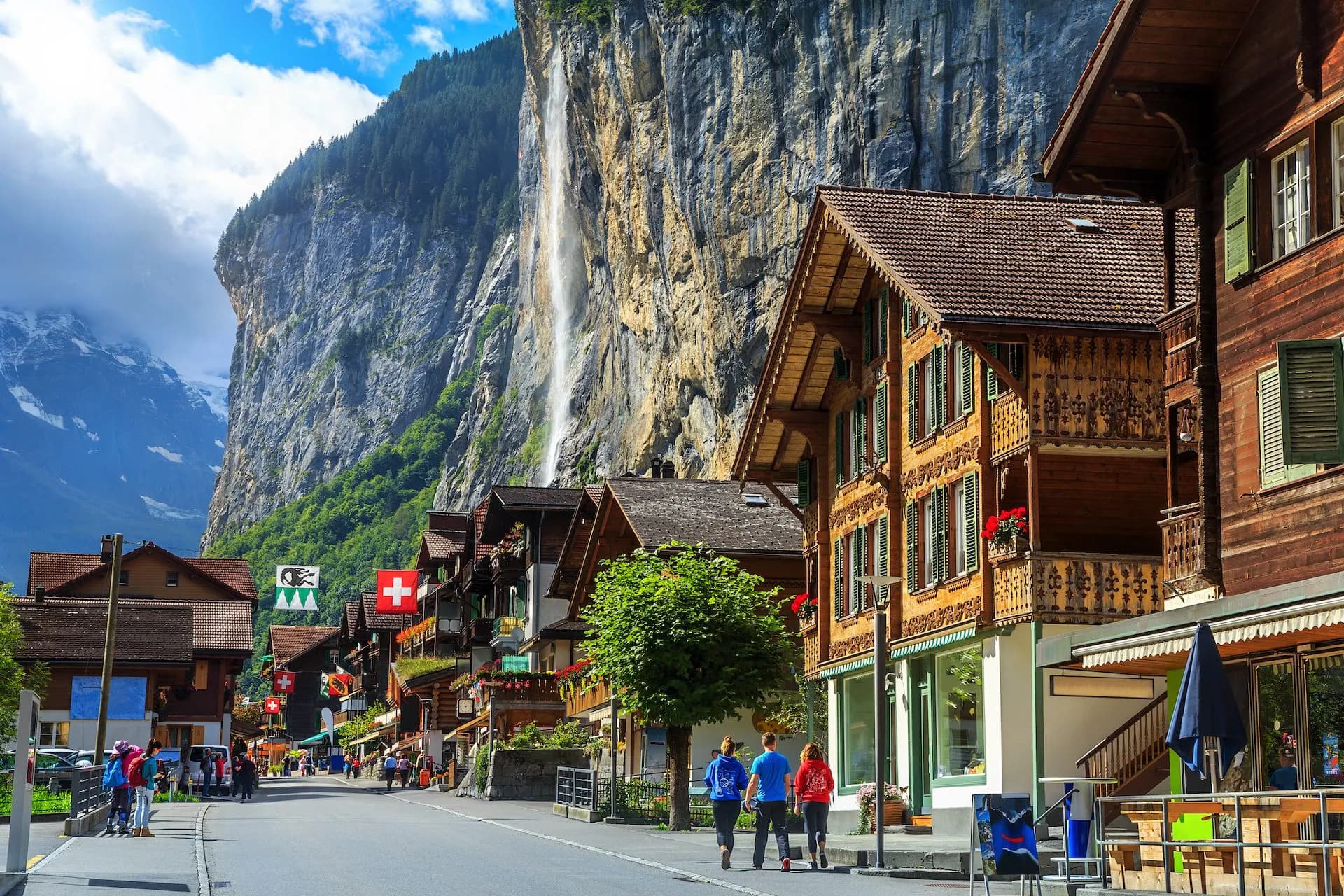 Lauterbrunnen street with traditional chalets, Staubbach Waterfall, and snow-capped mountains.