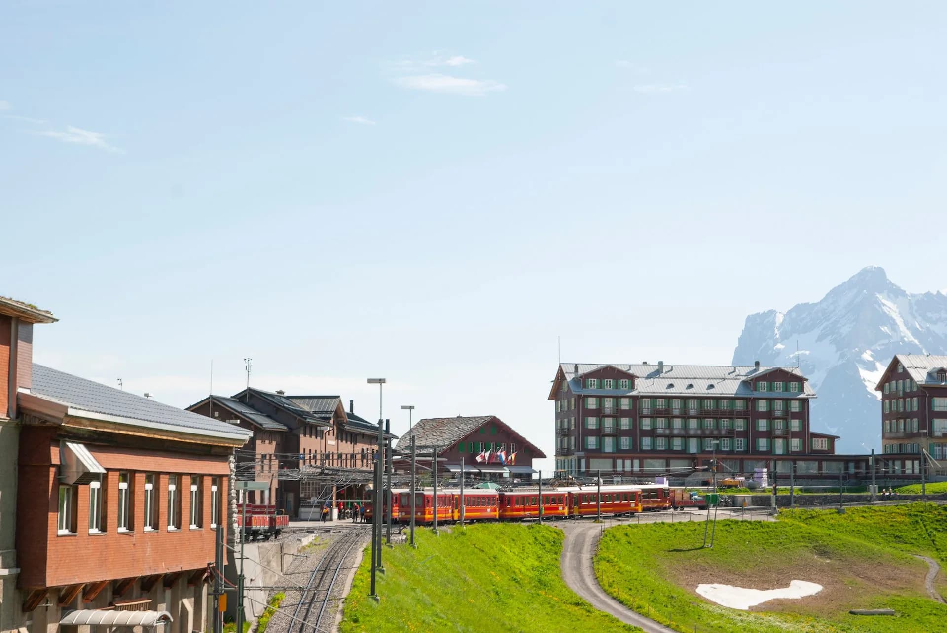 Kleine Scheidegg train station with red cogwheel train, green hillside, and snow-capped mountains.