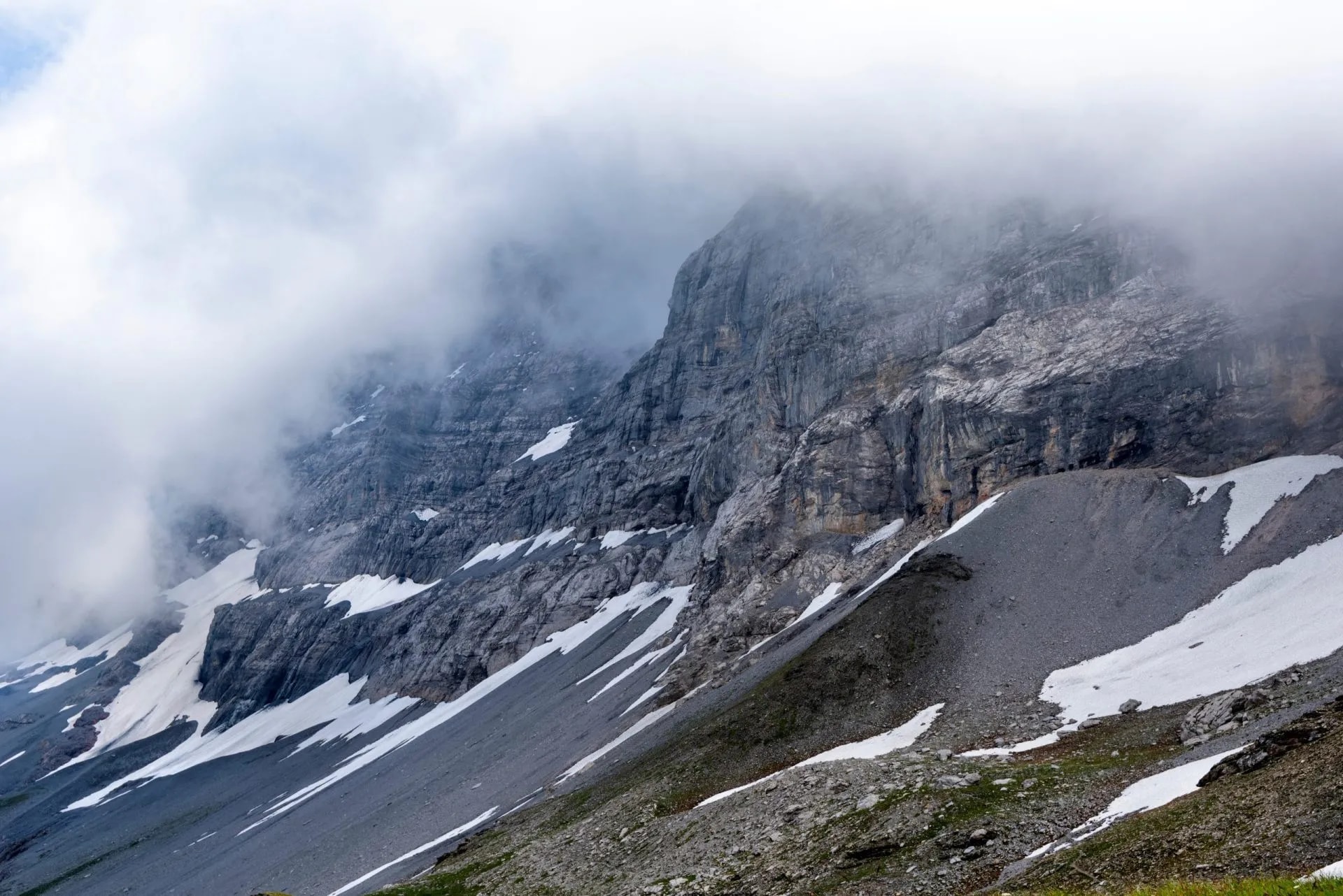 Steep gray mountain face with snow patches shrouded in low-hanging fog, Eiger North Face.