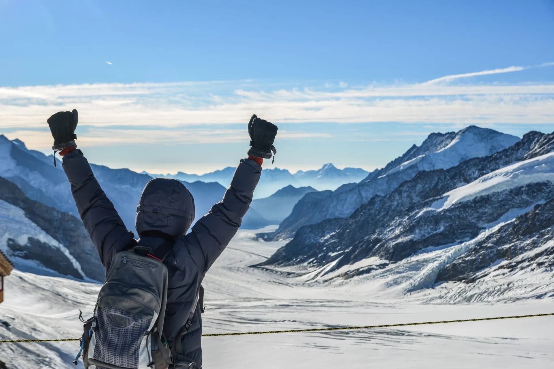 Hiker celebrating with arms raised overlooking the Aletsch Glacier and snow-covered Alpine mountains.